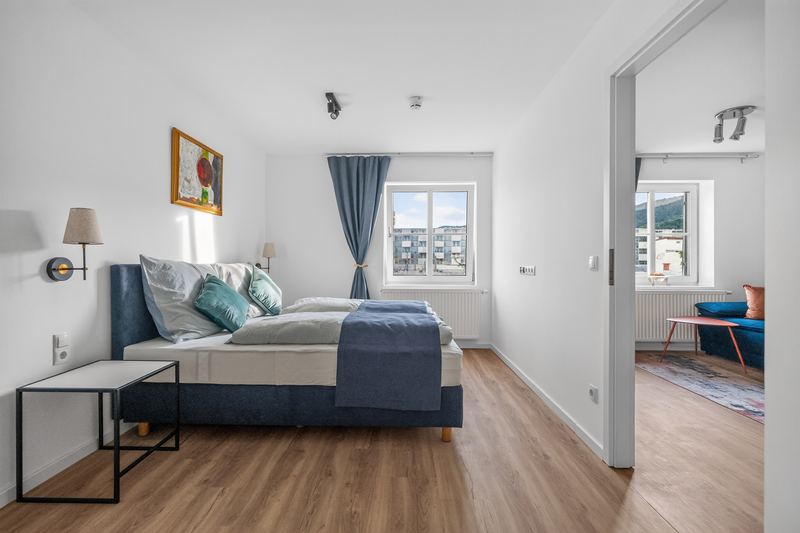 Top 6: bedroom with a double bed with blue headboard, bedside table and a view through the open door into the living room